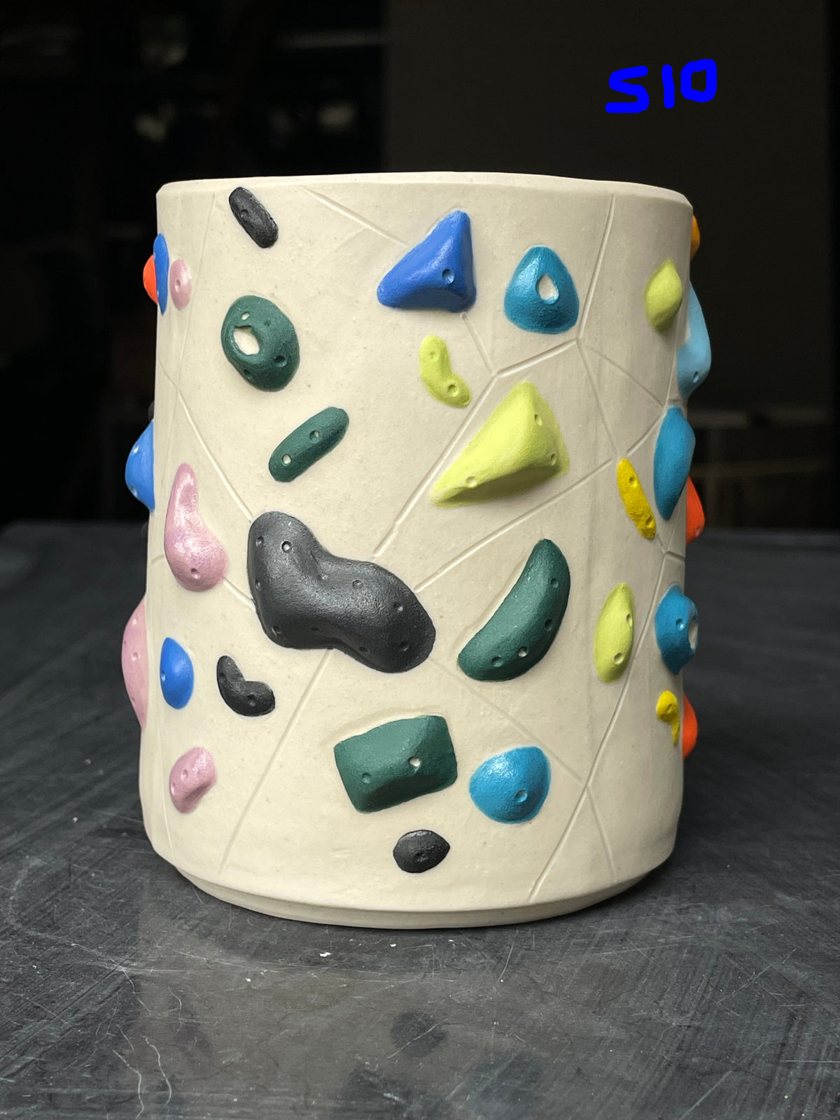 A cylindrical ceramic cup with a climbing-wall design, featuring a textured off-white base with multicolored climbing holds in blue, green, yellow, pink, orange, and black, displayed on a dark gray surface.