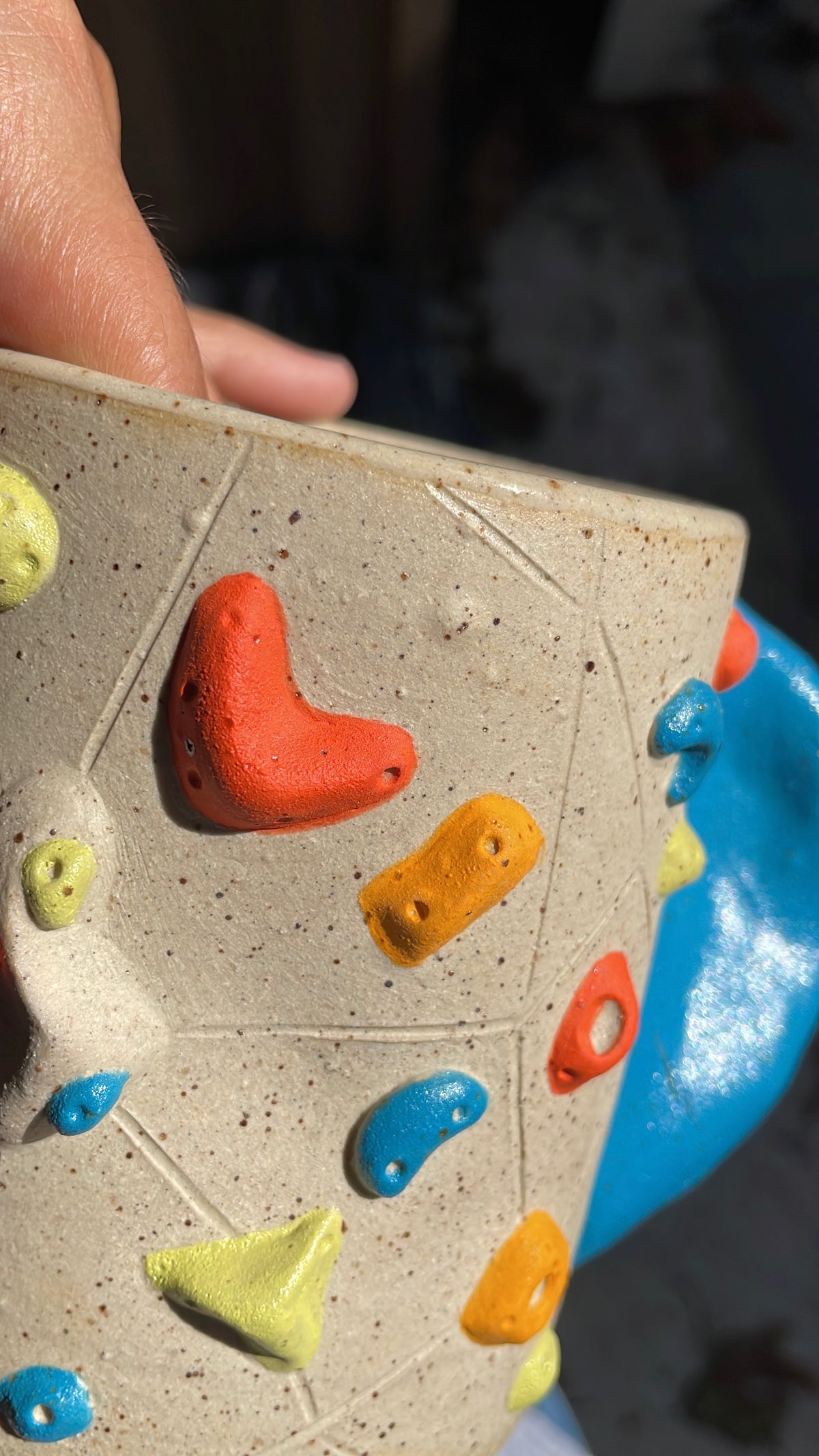 A close-up of a hand holding a speckled beige ceramic mug decorated with colorful climbing holds in orange, yellow, blue, and green, with a blue ceramic handle visible on the right.