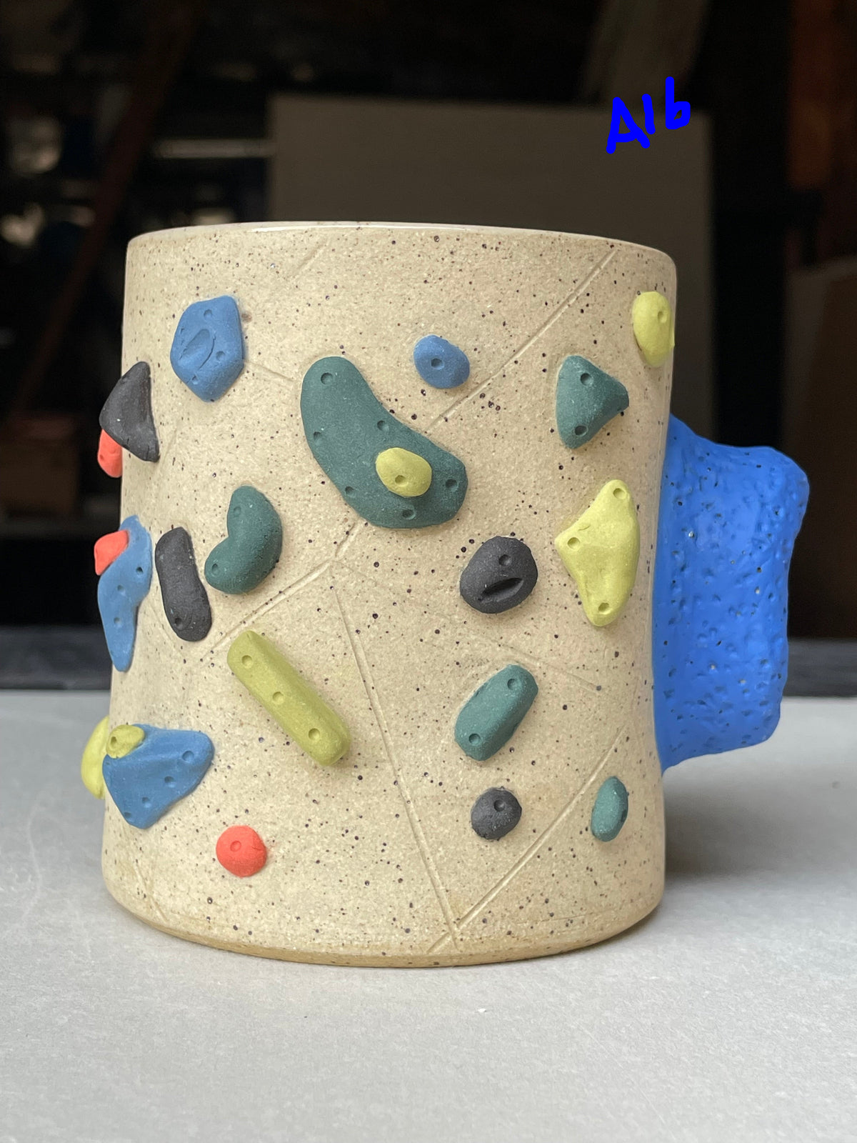 A speckled beige ceramic mug designed to look like a climbing wall, featuring multicolored climbing holds in blue, green, yellow, red, and gray, with a textured blue handle on the side.