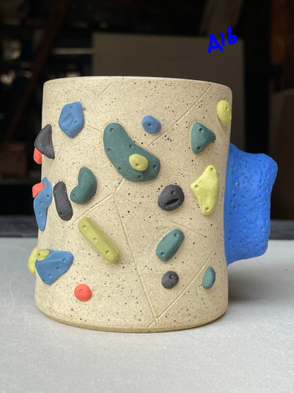 A speckled beige ceramic mug designed to look like a climbing wall, featuring multicolored climbing holds in blue, green, yellow, red, and gray, with a textured blue handle on the side.