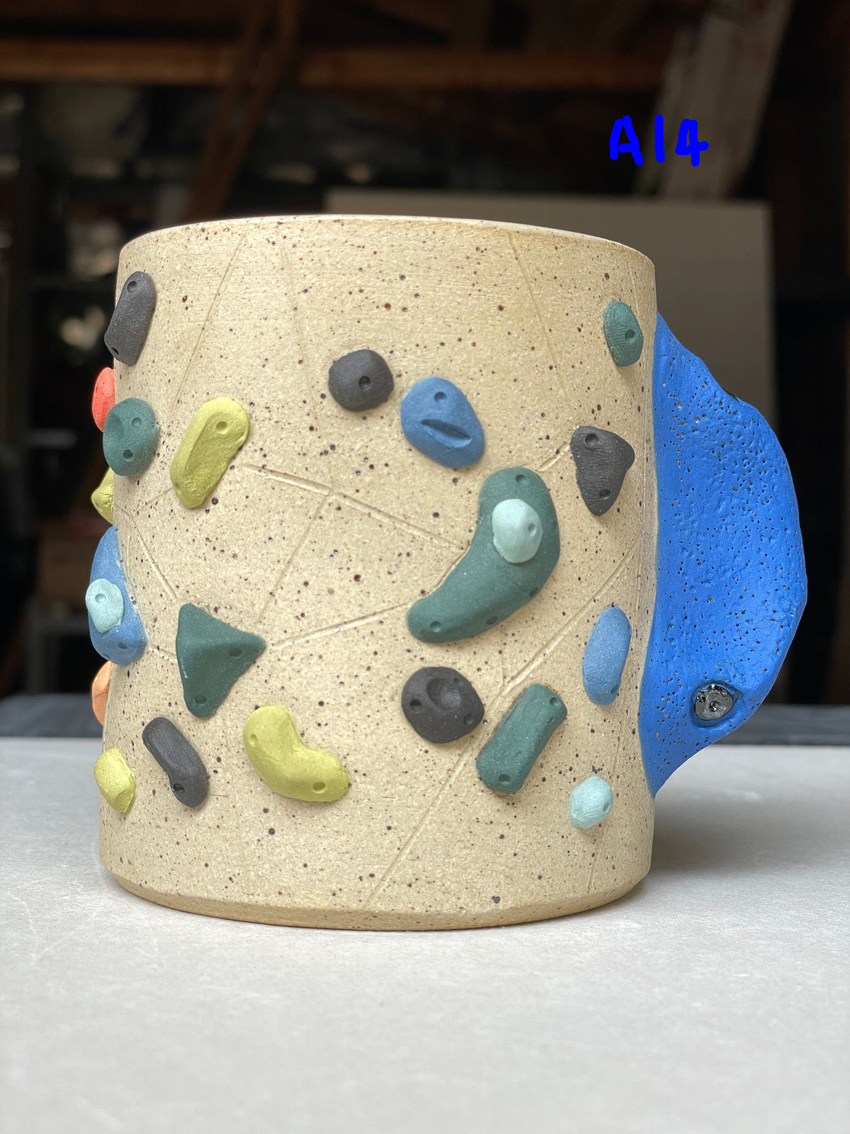 A handmade ceramic mug with a climbing-wall design, featuring a speckled beige body, colorful raised climbing holds, and a textured blue handle, set against a blurred workshop background.