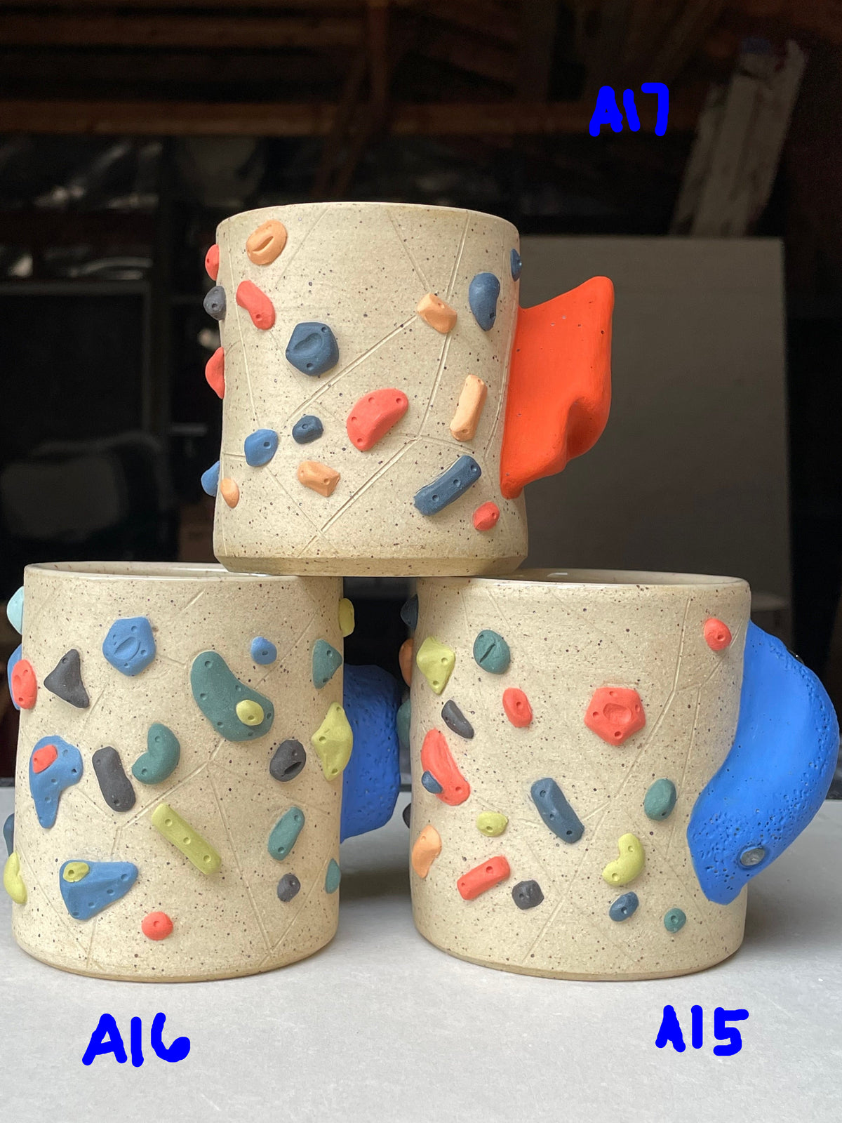 Three handmade ceramic mugs with climbing wall designs, stacked in a pyramid on a light gray surface. The mugs have a speckled cream-beige glaze and are decorated with colorful climbing holds in blue, orange, red, green, and yellow.